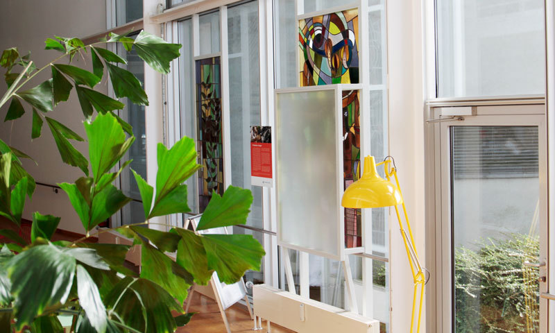 Eine Fotografie der Fensterbilder, im Vordergrund sind Blätter einer Pflanze zu sehen, rechts im Bild eine gelbe Stehlampe und links im Hintergrund leicht verschwommen, die Hussel-Fensterbilder, eines hinter einer Milchglasscheibe. 