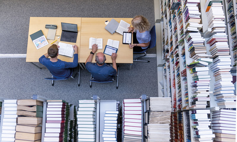 Vogelperspektive auf einen Tisch umgeben von Bücherregalen in der universitätsbibliothek. Am Tisch sitzen drei Personen mit Ihren Laptops, Tabletz und schreibutensilien.