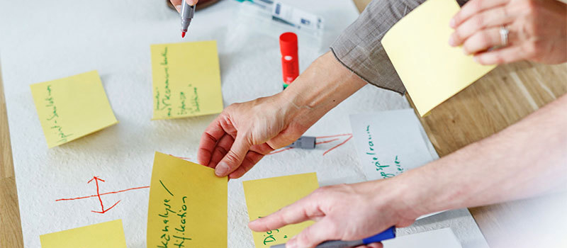 Schreibtisch mit Arbeitsutensilien zur Projektplanung. Im Bildausschnitt sichtbar Hände verschiedener Personen, die mit Post-its und Stiften eine Projektplanung auf Papier vornehmen.