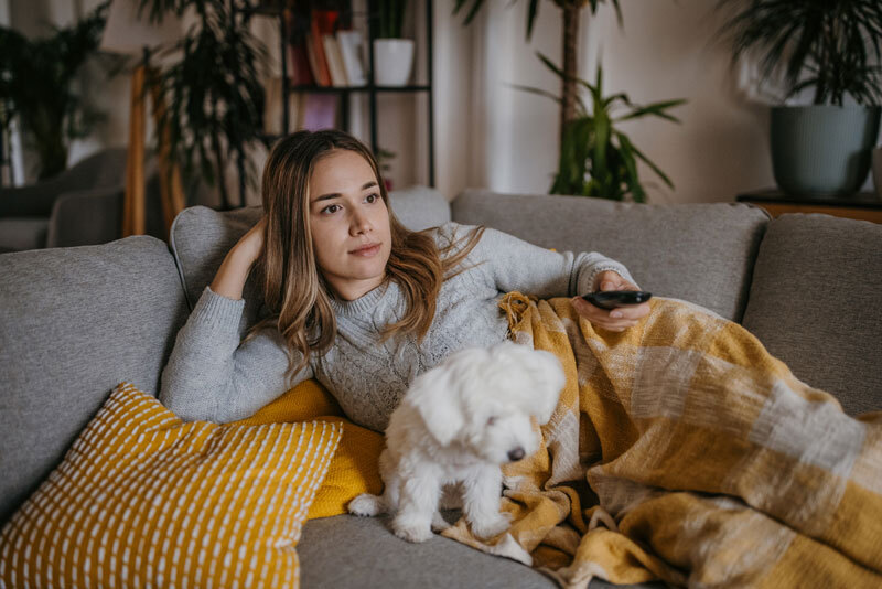 Eine junge Frau liegt mit ihrem Hund auf der Couch und schaut TV.