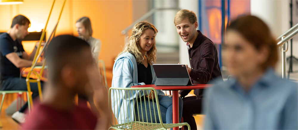 Two students working at a laptop computer in a communal space (other figures are blurred)