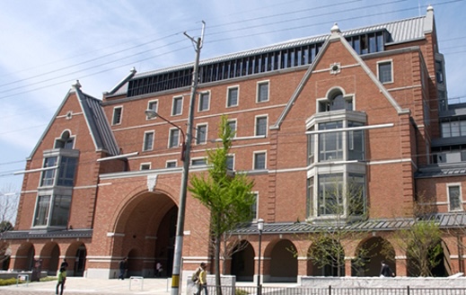 Ein Bild des Hauptgebäudes der Doshisha Law School. Es ist ein rotes Backsteingebäude mit einem grpßen Tor in der Mitte und zwei hervorstehenden Dachschrägen zu beiden Seiten.